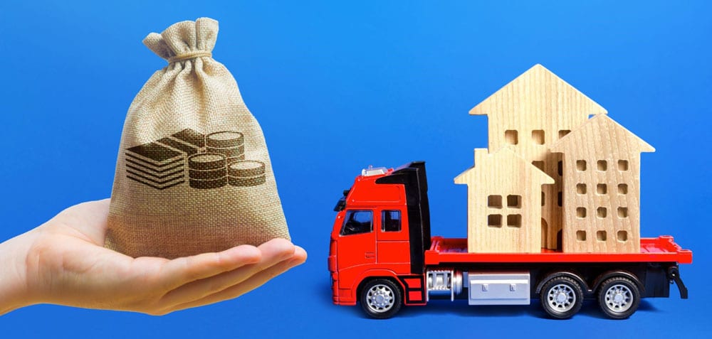 Hand holding bag of money next to toy truck with wood house figurines